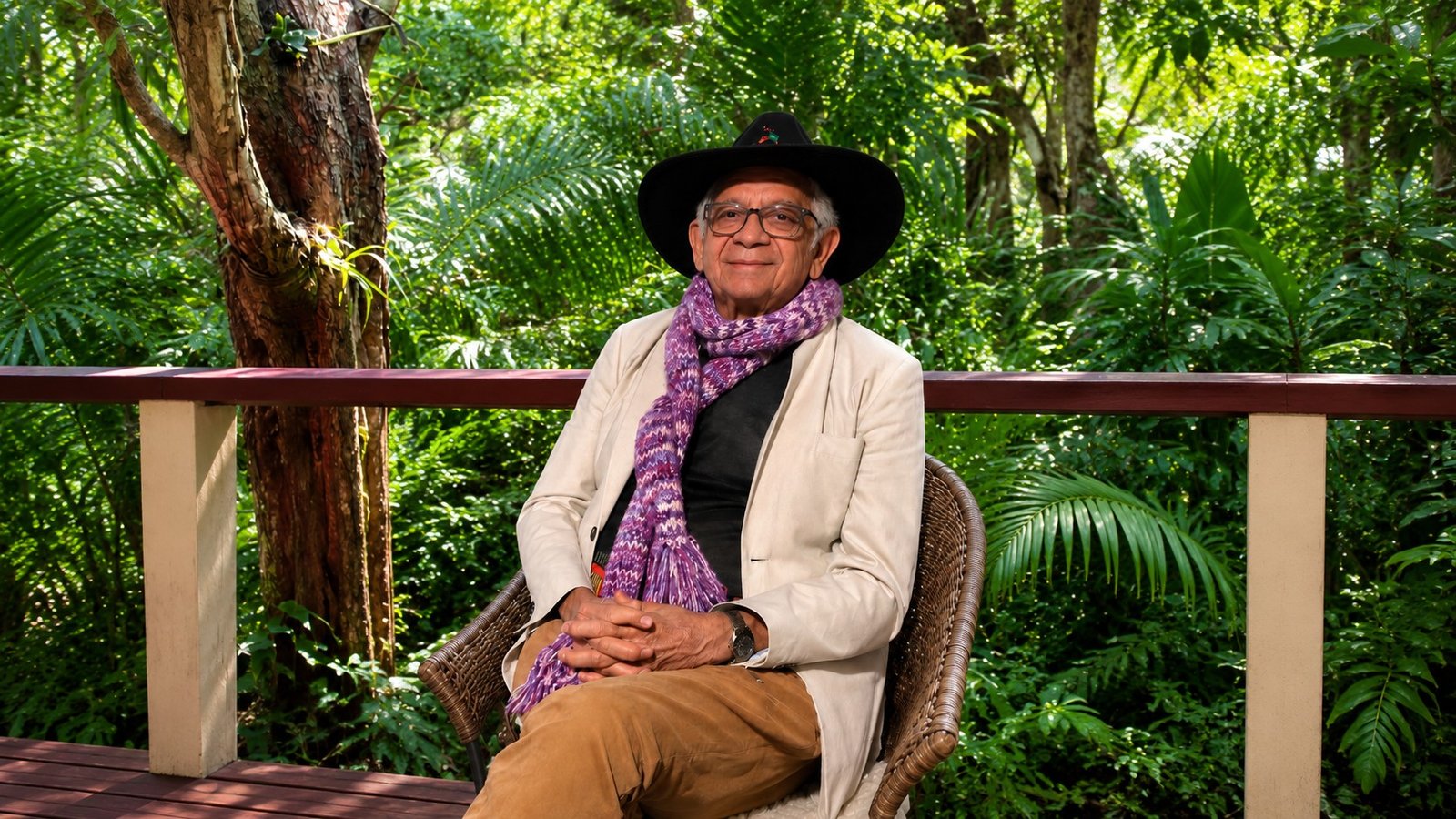 Uncle Bill Lemson seated on a back veranda in his hat and a purple scarf, green rainforest behind him