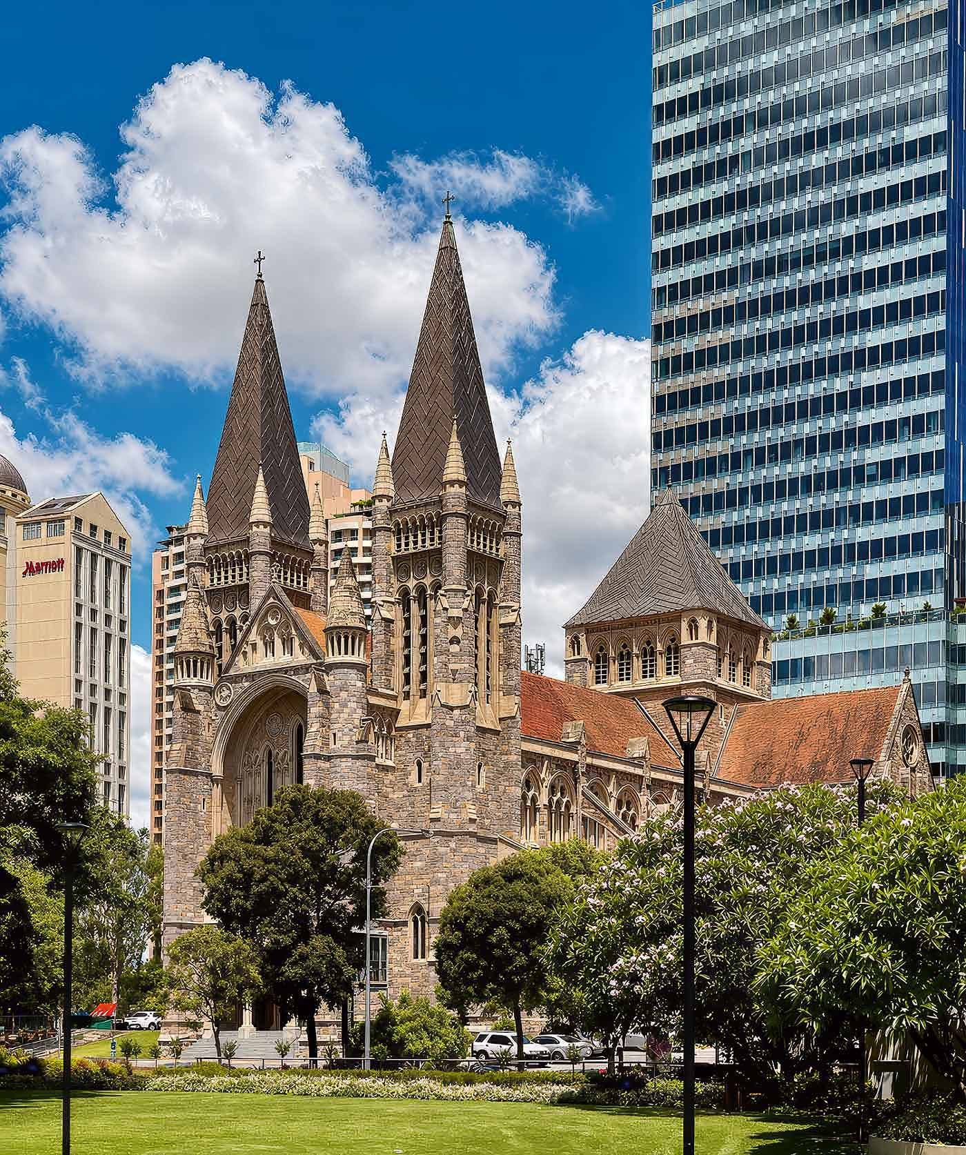 St John's Anglican Cathedral in Brisbane, twin sandstone spires rising against a blue sky, framed by trees and modern city buildings.