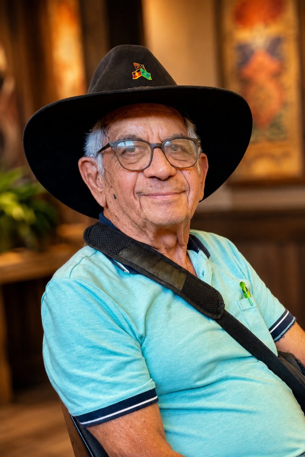Uncle Bill Lemson, smiling slightly toward the camera, wearing a wide-brimmed black hat with an enamelled Aboriginal flag badge, glasses, and a light blue polo shirt, with a bag strap visible across his shoulder. The background is a softly-lit interior space.