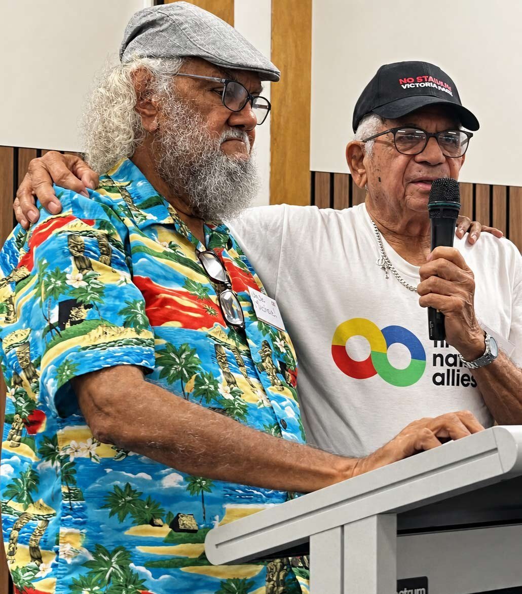 Uncle Bill Lemson, wearing a &lsquo;No Stadium Victoria Park&rsquo; cap and a First Nations Allies t-shirt, speaks into a handheld microphone at a podium. Beside him stands fellow Elder Uncle Michael Williams in a tropical-print shirt and grey flat cap, with one arm around Uncle Bill&rsquo;s shoulder.