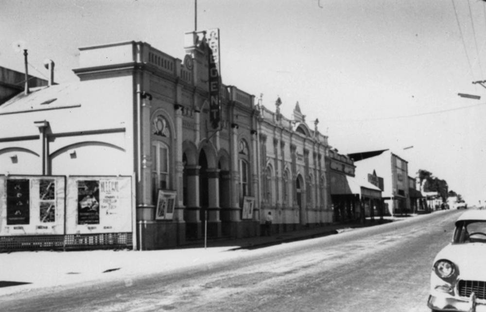 A wide main street in Charters Towers around 1950. The Regent Theatre stands on the corner under a tall vertical sign, shopfronts run away down the street beneath deep awnings, and a car of the period sits at the kerb.