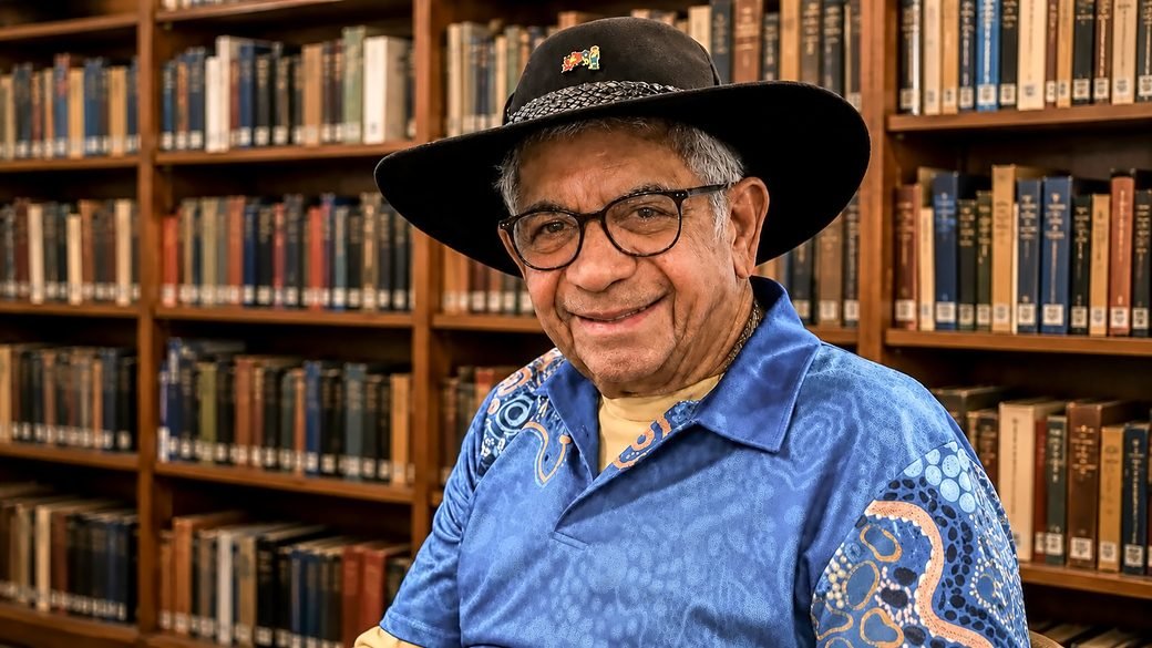 Uncle Bill Lemson seated in a library in his black hat and a blue shirt with First Nations designs, smiling toward the viewer.