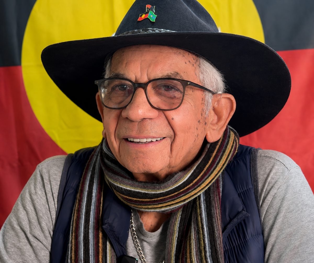 Uncle Bill Lemson smiling warmly in front of the Aboriginal Flag.