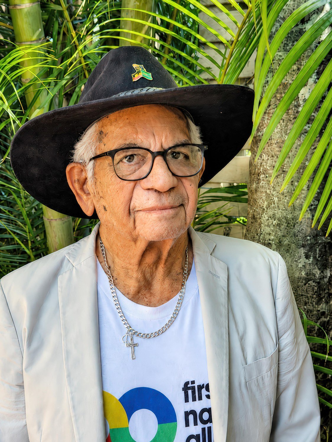 Uncle Bill Lemson, standing in a garden, wearing his hat with the Aboriginal Flag pin, a silver chain with a cross, and a First Nations Allies t-shirt under a cream blazer.