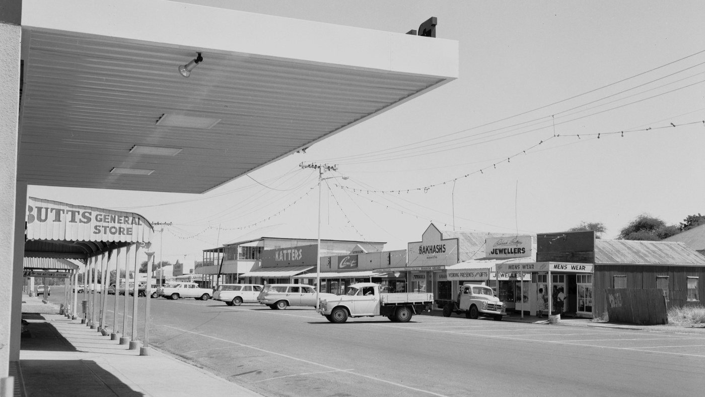 Ramsay Street, Cloncurry, in February 1966. Shopfronts under deep awnings, utes at the kerb, festive lights overhead.