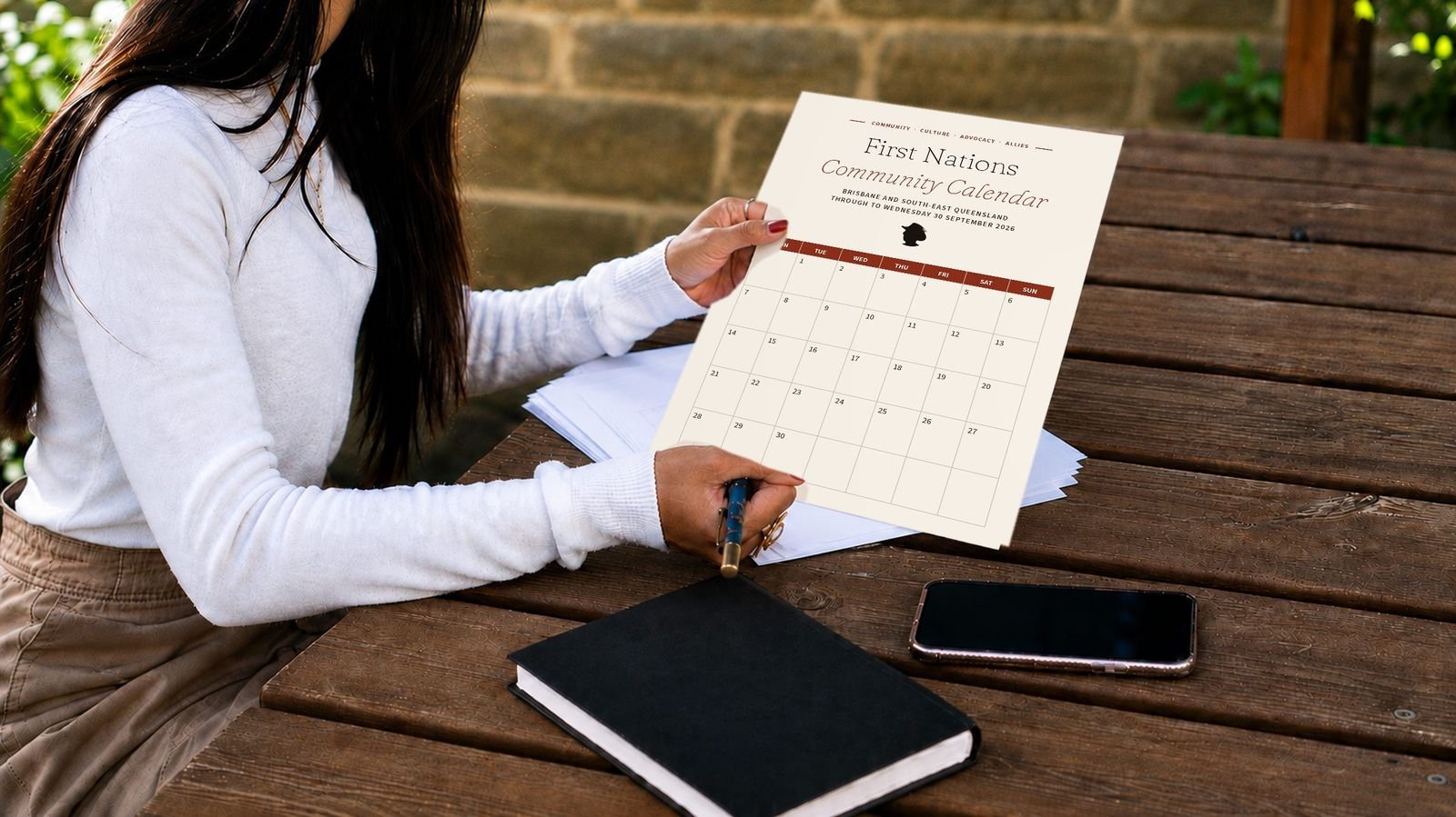 A woman at an outdoor table holding a printed page of the First Nations Community Calendar.