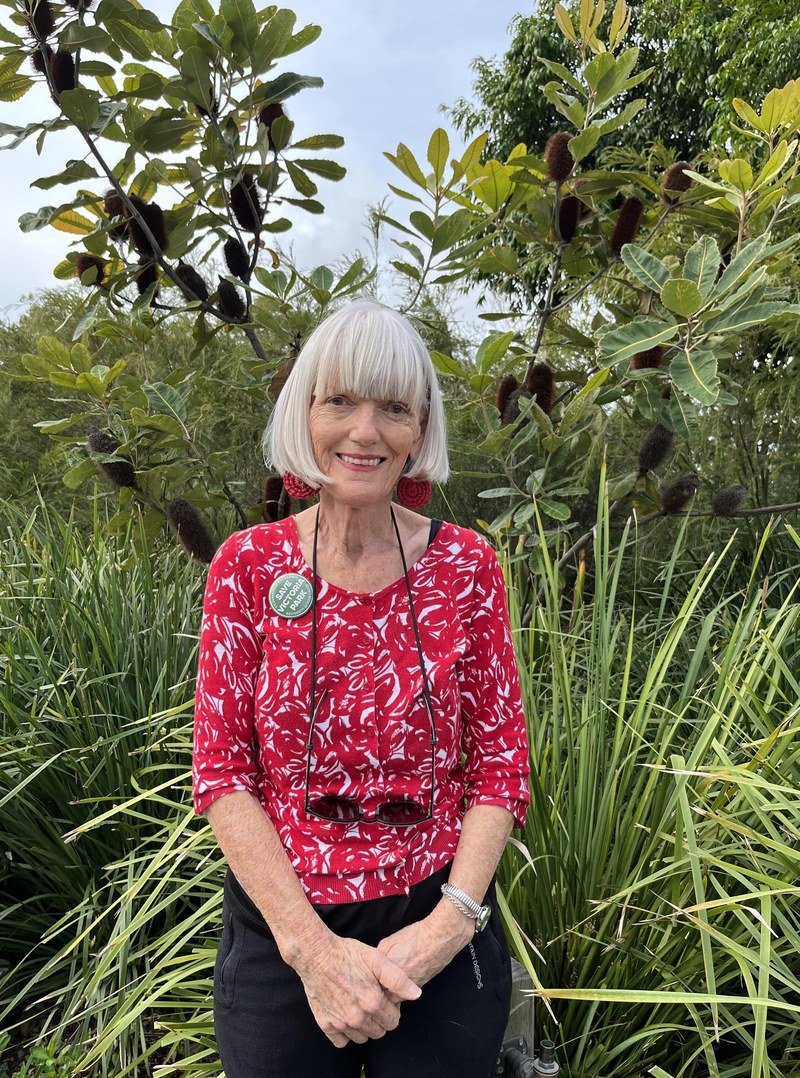 Linda Harnett, standing among banksia trees and native grasses at Barrambin (Victoria Park), wearing a red patterned cardigan and a 'Save Victoria Park' badge.