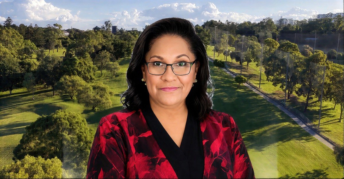 Aunty Sandra King OAM, Aboriginal Elder, with shoulder-length dark hair and glasses, wearing a black and red floral jacket, standing before the green parkland of Barrambin.