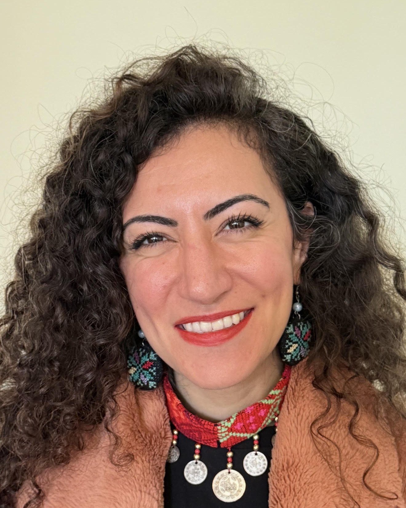 Remah Naji, a woman with long dark curly hair, smiling warmly at the camera, wearing embroidered Palestinian tatreez earrings and a traditional coin necklace over a soft brown coat.