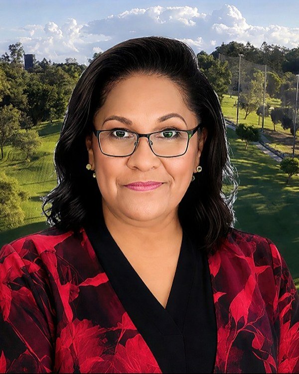 Aunty Sandra King OAM, Aboriginal Elder, with shoulder-length dark hair and glasses, wearing a black and red floral jacket, with the green parkland of Barrambin behind her.