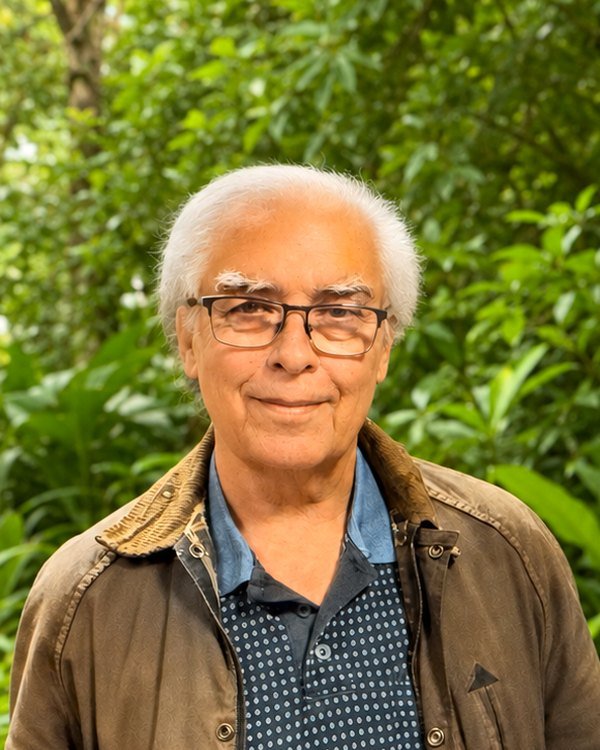 Colin Thomas, a man with white hair and glasses in a brown jacket, in front of green bushland.